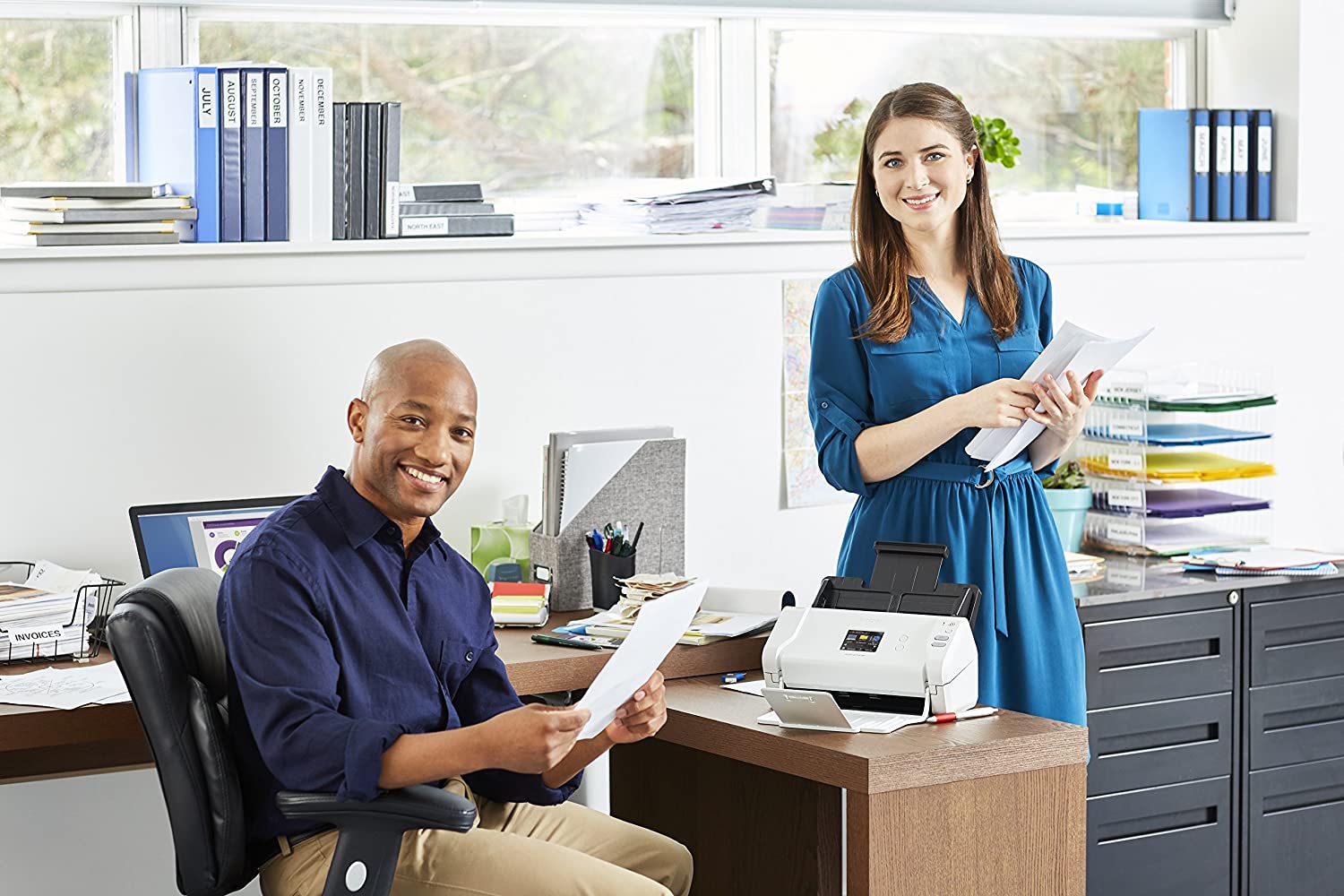 man sitting at a work desk and woman standing, both are smiling and holding sheets of paper